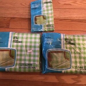 May include: Three packages of no-iron muslin bedding in a green and white gingham check pattern. The set includes one flat twin sheet, one fitted twin sheet, and two standard pillowcases, all sealed in original plastic packaging with blue accents.