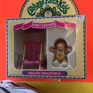 May include: A boxed Cabbage Patch Kids Deluxe Miniatures set. The box features a doll with red hair and a yellow outfit, along with a miniature pink and white swing set. The box reads "First Edition" and "For Ages 3 and Up."