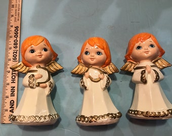 May include: Three ceramic angel figurines with orange hair, white dresses, and gold accents. The angels are holding a harp, a book, and praying hands.