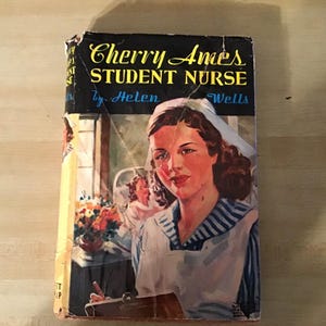 May include: Vintage book cover for "Cherry Ames, Student Nurse" by Helen Wells. The cover art features a woman in a nurse's uniform with a white cap and blue striped top. The title is in yellow and the author's name is in blue.