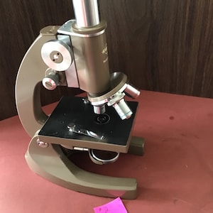 May include: A vintage brown microscope with a silver metal arm and a black stage. The microscope has a single eyepiece and a rotating nosepiece with two objective lenses. The microscope is sitting on a red surface.
