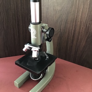 May include: A green and black microscope with a black stage and a silver and black body. The microscope has a black knob on the side and a black and silver knob on the top. The microscope is sitting on a red surface.