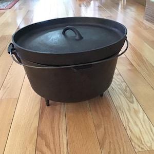 May include: A dark brown cast iron Dutch oven with a lid and handle. The pot has three short legs and a wire handle. The lid has a loop handle. This cookware is ideal for camping and outdoor cooking.