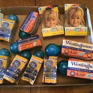 May include: A collection of vintage photoflash bulbs and flash cubes in a cardboard box. The bulbs are blue and have a sparkly finish. The boxes are yellow, orange, and white with black text. The text on the boxes reads "Westinghouse Photoflash Bulb", "GE Lamps Photoflash", and "Sylvania Flashcubes".