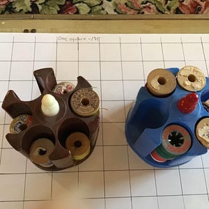 May include: Two thread spool holders, one brown and one blue, each holding several spools of thread. The brown holder has a white knob, and the blue holder has a red knob. The holders are on a grid.