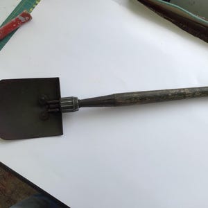 May include: A folding entrenching tool featuring a dark metal spade head with a pointed tip and a weathered wooden handle. The spade is connected to the handle by a metal collar mechanism, showing signs of wear and use on the wood surface.