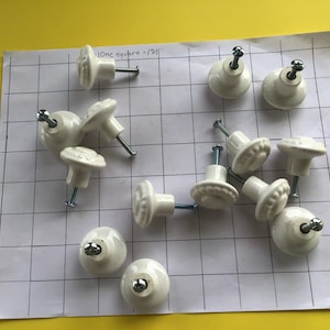 May include: A collection of white ceramic cabinet knobs with silver-toned hardware. The knobs are arranged on a grid-lined paper, with the text "1one square = 1/8"" visible. The knobs have a decorative, possibly floral, design.