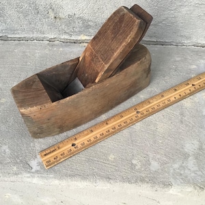 May include: A vintage wooden hand plane with a ruler. The plane is made of aged wood and has a rectangular body with a blade. The ruler is made of wood and is marked with inch measurements. The items are on a gray surface.