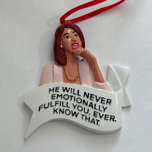 May include: White ceramic Christmas ornament featuring a woman with red hair and a pink shirt. The ornament has a speech bubble that reads "He will never emotionally fulfill you. Ever. Know that."