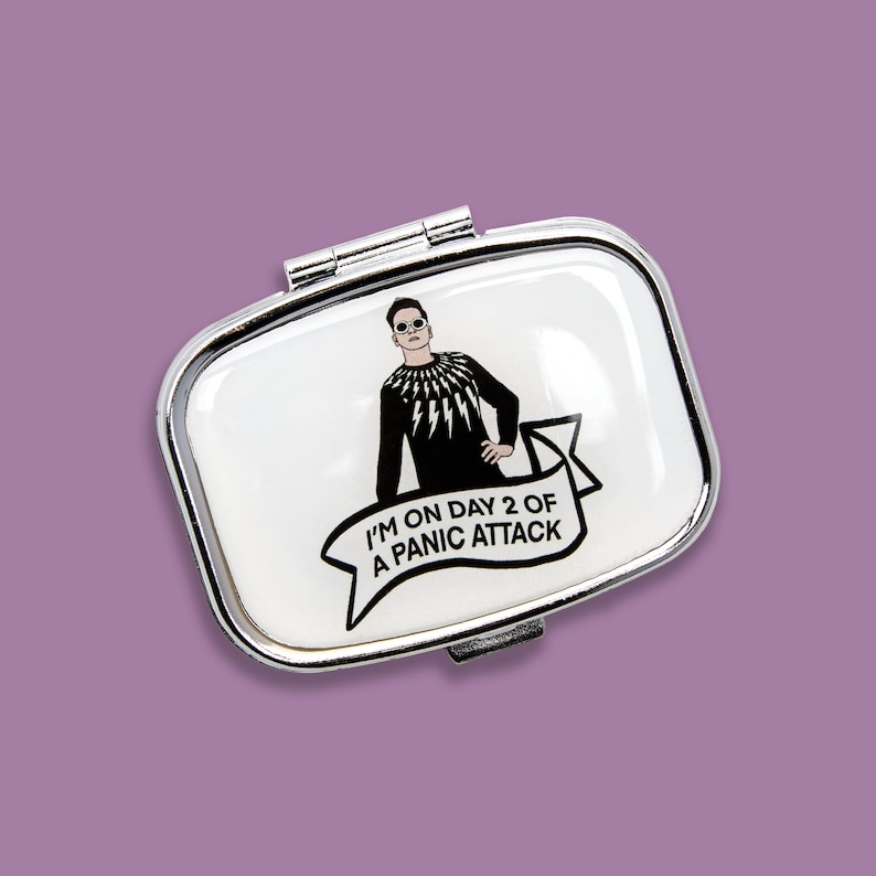 K&ouml;nnte beinhalten: Eine silberne Pillendose mit wei&szlig;em Hintergrund und einer Schwarzwei&szlig;illustration einer Person, die ein schwarzes Hemd mit einem Blitz-Design tr&auml;gt. Der Text auf dem Banner lautet "Ich bin im zweiten Tag einer Panikattacke".