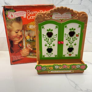 May include: A Strawberryland Miniatures Berry Patch Garden toy display case with a tan frame, white doors featuring green trim and strawberry cutouts, and a floral base with the text Life is Berry Delicious, accompanied by its original cardboard box.