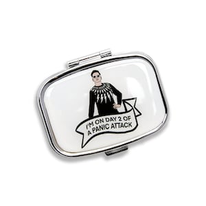 K&ouml;nnte beinhalten: Eine silberne Pillendose mit wei&szlig;em Hintergrund und einer Schwarzwei&szlig;illustration einer Person mit Sonnenbrille und einem schwarzen Shirt mit Blitzen. Der Text "I'm on day 2 of a panic attack" ist auf einem wei&szlig;en Banner gedruckt.