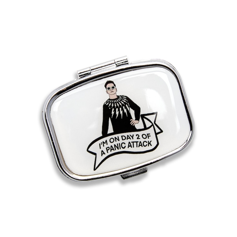 K&ouml;nnte beinhalten: Eine silberne Pillendose mit wei&szlig;em Hintergrund und einer Schwarzwei&szlig;illustration einer Person mit Sonnenbrille und einem schwarzen Shirt mit Blitzen. Der Text "I'm on day 2 of a panic attack" ist auf einem wei&szlig;en Banner gedruckt.