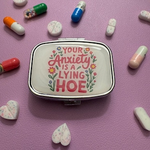 May include: A silver pill case with the text "YOUR Anxiety IS A LYING HOE" surrounded by floral designs. Various pills and capsules in different colors are scattered around the case on a purple surface.