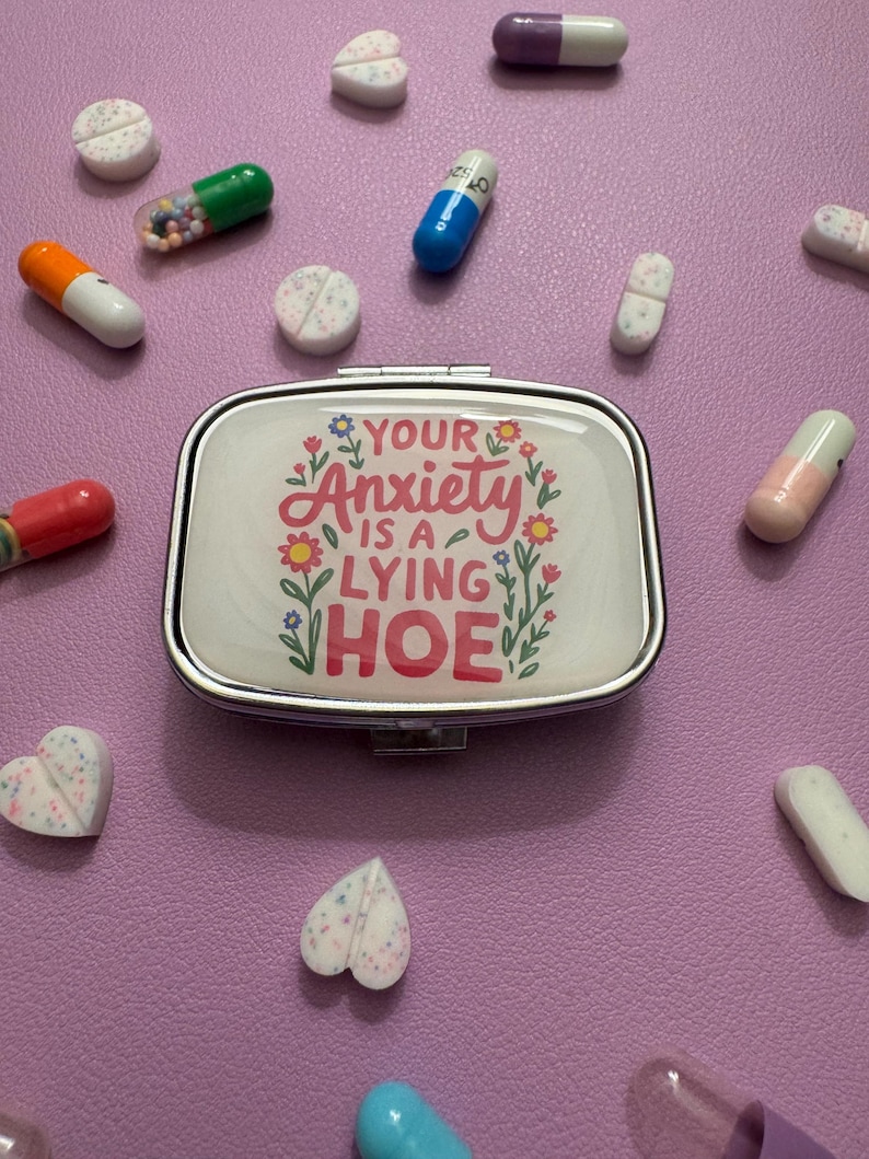May include: A silver pill case with the text "YOUR Anxiety IS A LYING HOE" surrounded by floral designs. Various pills and capsules in different colors are scattered around the case on a purple surface.