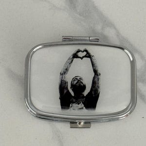 May include: A silver compact mirror with a black and white image of a person making a heart shape with their hands.