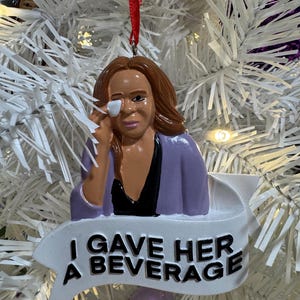 May include: A Christmas tree ornament featuring a woman with brown hair and a purple jacket, holding her eye. The ornament has the text "I GAVE HER A BEVERAGE" on a white banner. The ornament is hanging on a white Christmas tree.