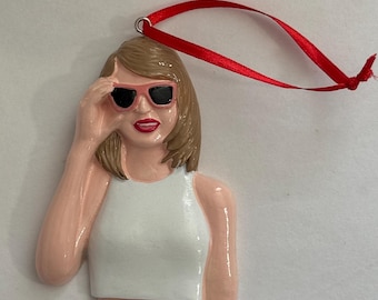 May include: A pink and white ceramic ornament of a woman wearing sunglasses and a white tank top. The ornament has a red ribbon attached to the top.