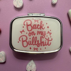 May include: A silver pill case with the phrase "Back on my Bullshit" in pink script. The case is surrounded by various pills and capsules on a purple surface. The text is decorated with bows and stars.