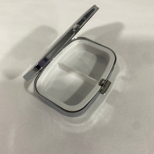 May include: A small, rectangular pill box with a hinged lid. The box is silver-colored with a white interior divided into two compartments. The lid is partially open, revealing the inside. The box is designed for storing medication or small items.