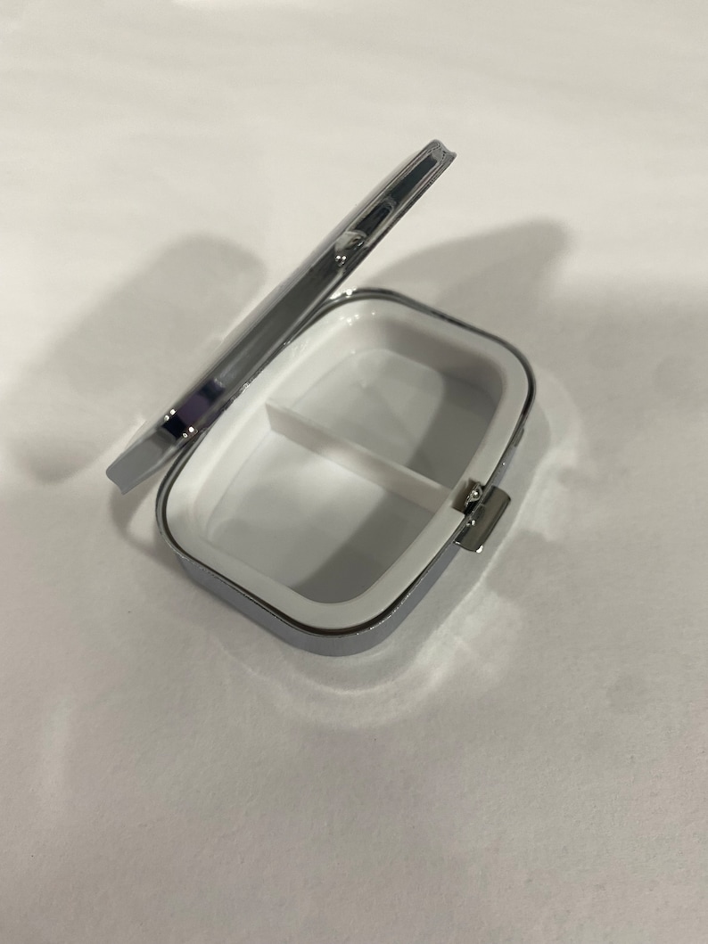 May include: A small, rectangular pill box with a hinged lid. The box is silver-colored with a white interior divided into two compartments. The lid is partially open, revealing the inside. The box is designed for storing medication or small items.