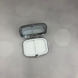 May include: A silver-colored pill box, open to reveal two white compartments. The box has a rectangular shape with rounded corners and a hinged lid. The interior is divided into two sections.