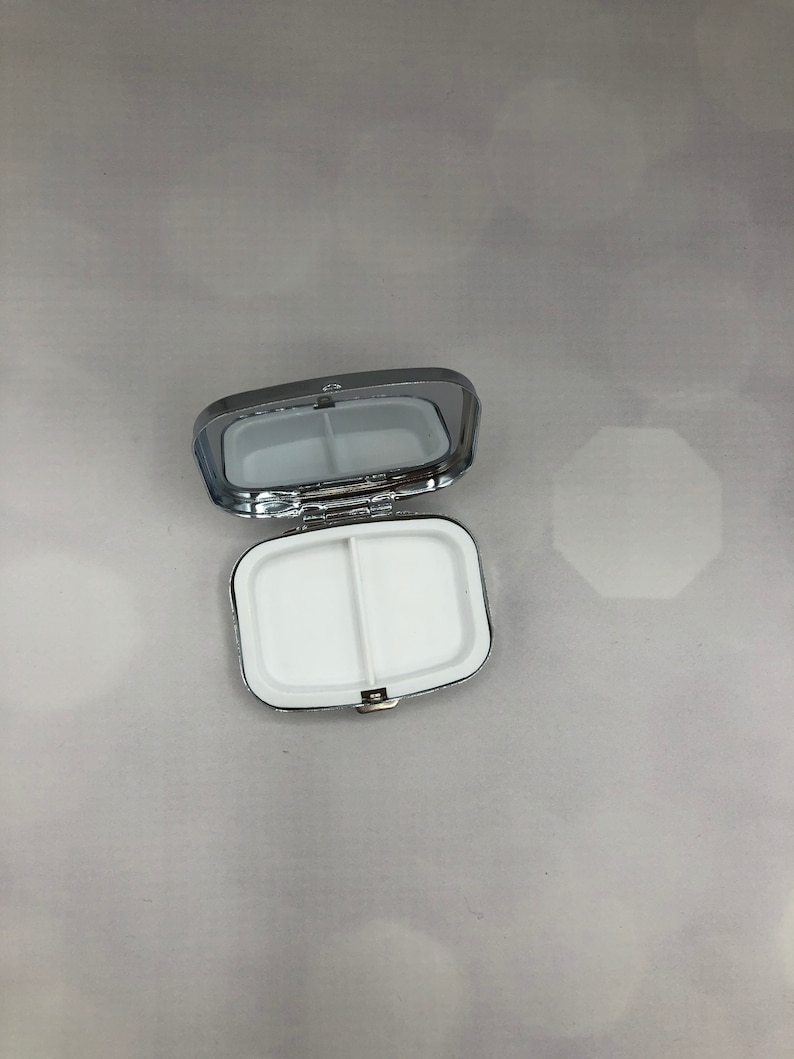 May include: A silver-colored pill box, open to reveal two white compartments. The box has a rectangular shape with rounded corners and a hinged lid. The interior is divided into two sections.