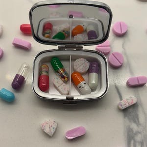 May include: A small, silver pill box is open, revealing various colorful pills and capsules. The pills are scattered around the box on a white surface. The box has a mirrored lid. The pills are in various shapes and colors, including pink, purple, orange, and green.