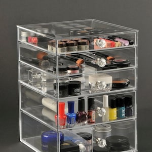 May include: Clear acrylic makeup organizer with six drawers. The drawers are filled with various makeup products, including lipsticks, eyeshadows, nail polish, and brushes.