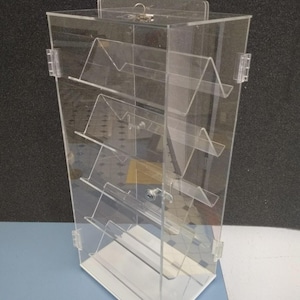 May include: Clear acrylic display case with multiple angled shelves. The case has a white base and a hinged door with a lock. The display is designed to showcase items and is suitable for retail or personal use.