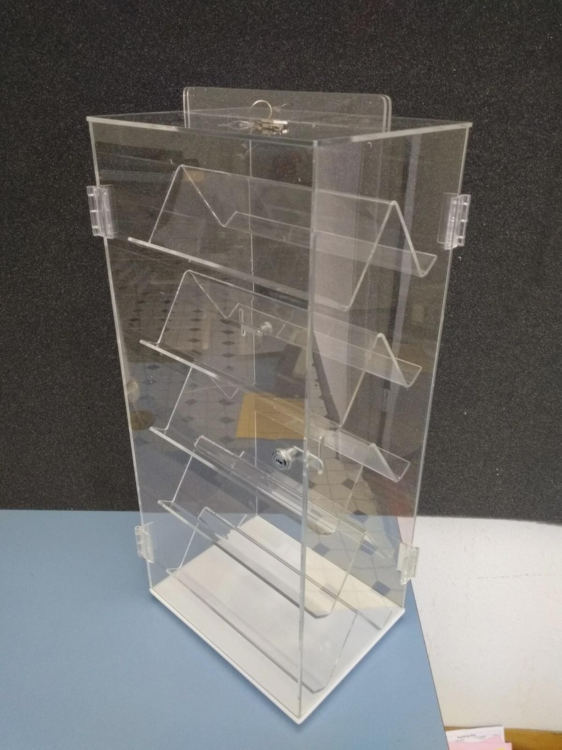 May include: Clear acrylic display case with multiple angled shelves. The case has a white base and a hinged door with a lock. The display is designed to showcase items and is suitable for retail or personal use.