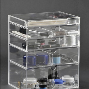 May include: Clear acrylic organizer with five drawers for storing makeup and other small items. The drawers are filled with various cosmetics, including nail polish, eye shadow, and lip gloss.