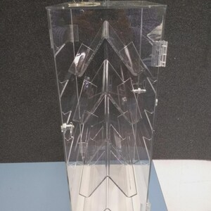 May include: A clear acrylic display case with a hinged door and a lock. The interior features multiple angled shelves, creating a tiered presentation space. The case is designed to showcase items and is set against a neutral background.