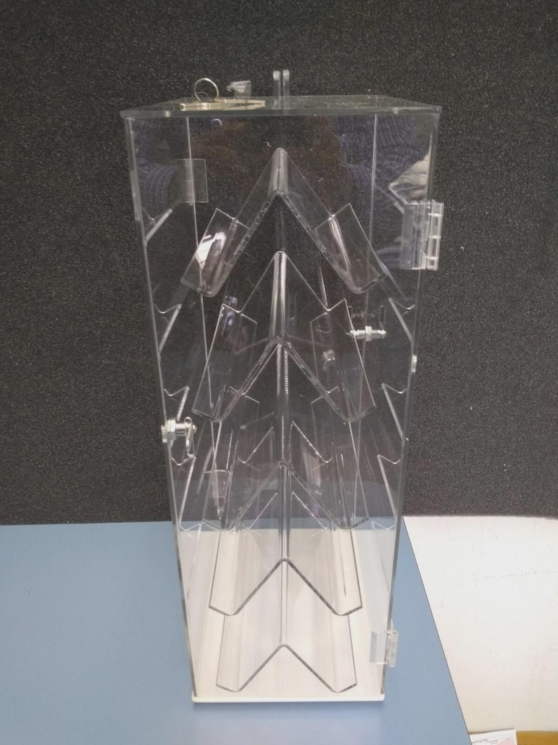 May include: A clear acrylic display case with a hinged door and a lock. The interior features multiple angled shelves, creating a tiered presentation space. The case is designed to showcase items and is set against a neutral background.