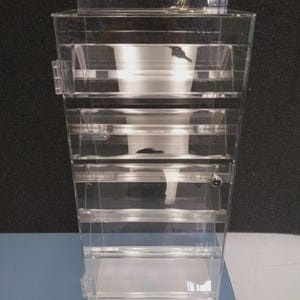 May include: A clear, multi-tiered acrylic display case with a hinged lid and small metal latches. The case has several horizontal shelves and a rectangular base. The interior features a funnel-shaped design. The background is a dark gray.