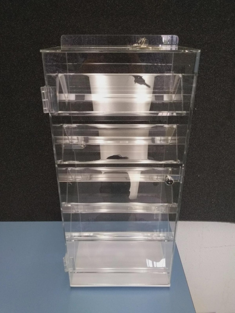 May include: A clear, multi-tiered acrylic display case with a hinged lid and small metal latches. The case has several horizontal shelves and a rectangular base. The interior features a funnel-shaped design. The background is a dark gray.