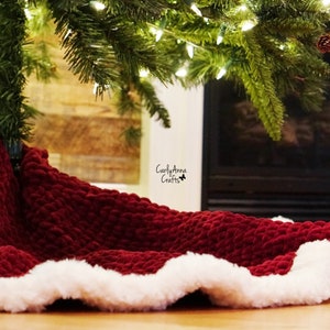 May include: A red velvet Christmas tree skirt with a white faux fur trim. The skirt is placed under a Christmas tree in front of a fireplace.