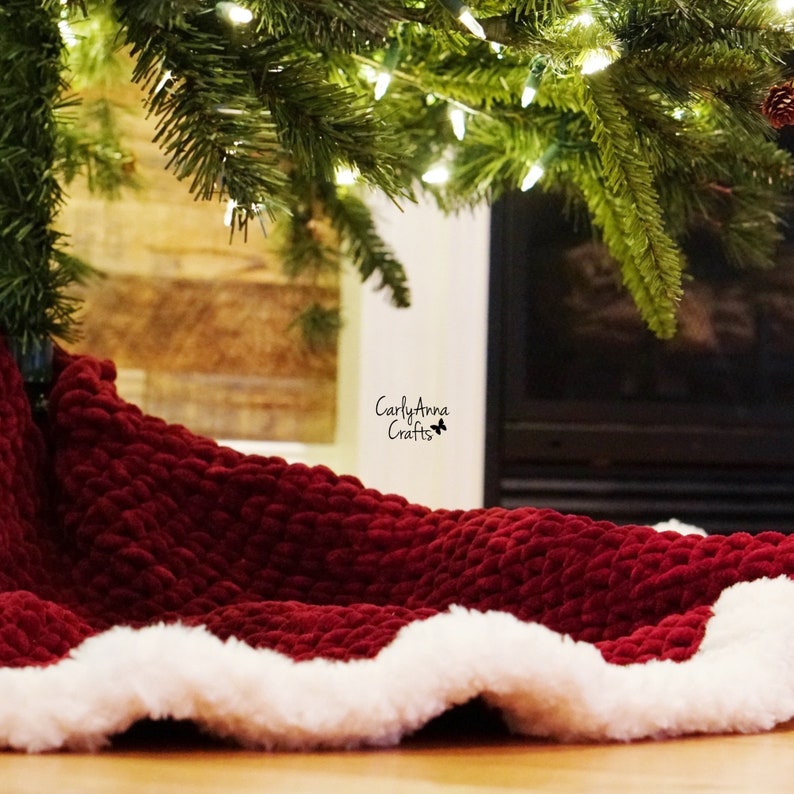 May include: A red velvet Christmas tree skirt with a white faux fur trim. The skirt is placed under a Christmas tree in front of a fireplace.