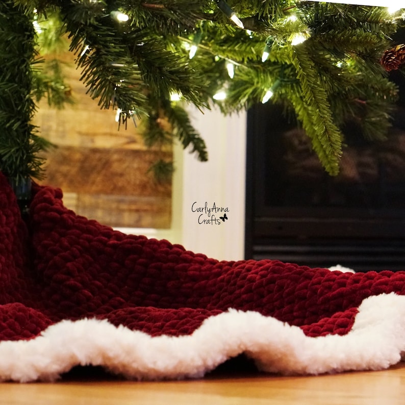 May include: A red velvet Christmas tree skirt with a white faux fur trim. The skirt is under a Christmas tree with lights and is in front of a fireplace.