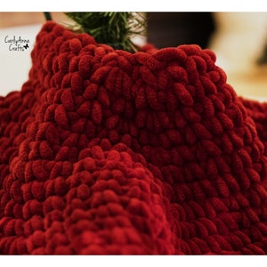 May include: Close-up of a deep red crocheted blanket with a soft, fuzzy texture. The blanket is made with thick yarn and has a unique, textured pattern.