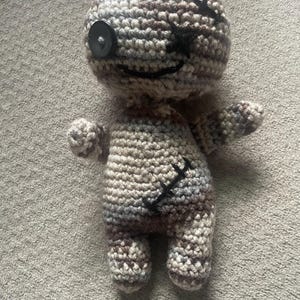 Könnte beinhalten: Handgefertigte Häkelpuppe in Grau-, Braun- und Cremetönen. Die Puppe hat ein Knopfauge und schwarze, aufgestickte Details für Mund und Körper. Die Arme und Beine sind klein und abgerundet, was ihr ein skurriles Aussehen verleiht.