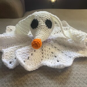 May include: A handmade crochet blanket featuring a white ghost dog with black eyes and an orange nose. The blanket has a scalloped edge and is made of white yarn. The dog's head is three-dimensional.