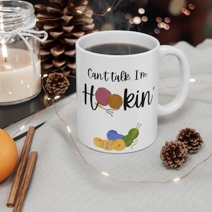 Puede incluir: Taza de cerámica blanca con el texto "Can't talk. I'm Hookin'" e ilustraciones de ovillos de lana y ganchillos. La taza tiene un asa curva y está llena de un líquido oscuro. Elementos decorativos: piñas y una vela encendida.