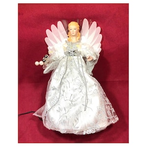 May include: A Christmas tree topper in the shape of an angel. The angel has blonde hair, a white dress with silver accents, and white feathered wings. She is holding a silver beaded strand and is set against a red background.