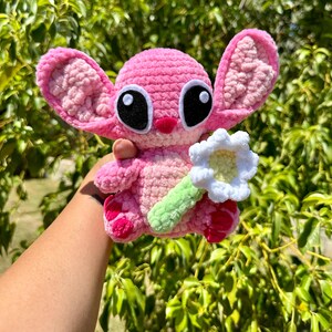 May include: Pink crocheted plush toy with large black eyes and a white crocheted flower. The toy is holding the flower in its right hand.