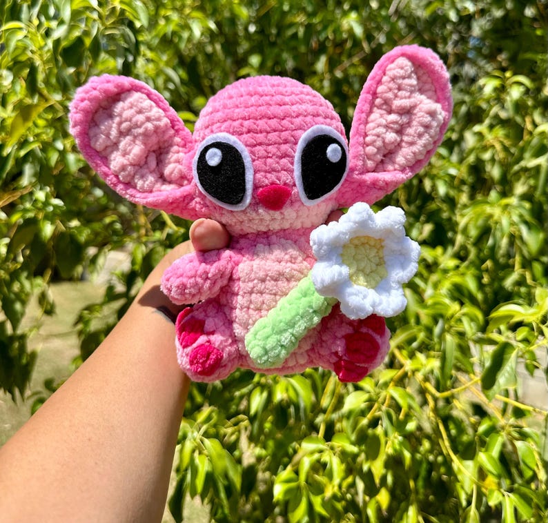 May include: Pink crocheted plush toy with large black eyes and a white crocheted flower. The toy is holding the flower in its right hand.
