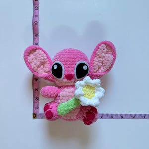 May include: Pink crocheted plush toy with large black eyes and a white crocheted flower. The toy is holding a green crocheted stem.