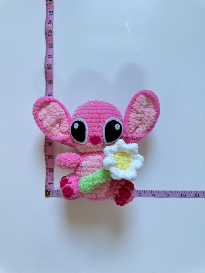 May include: Pink crocheted plush toy with large black eyes and a white crocheted flower. The toy is holding a green crocheted stem.