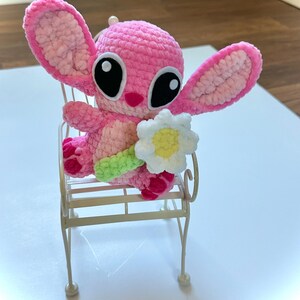 May include: A pink crocheted Stitch plush toy holding a white crocheted flower. The toy is sitting on a white metal chair.