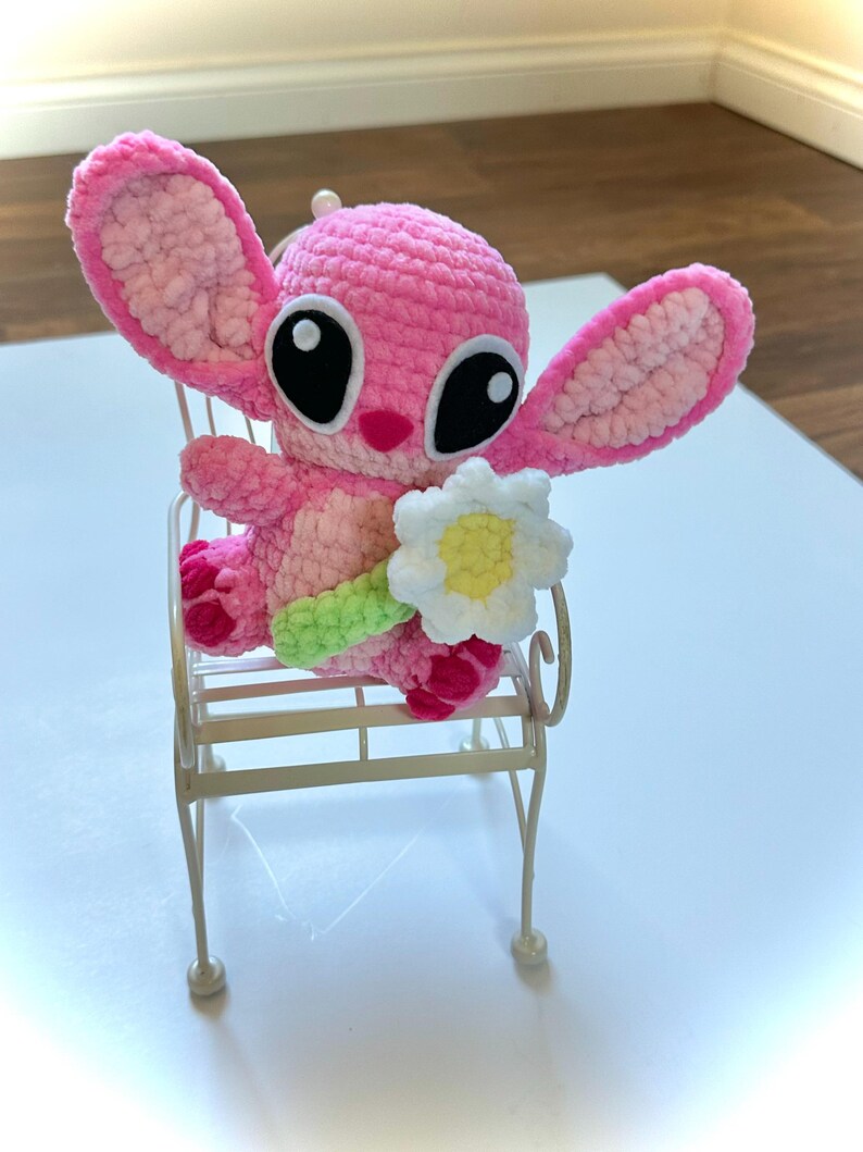 May include: A pink crocheted Stitch plush toy holding a white crocheted flower. The toy is sitting on a white metal chair.